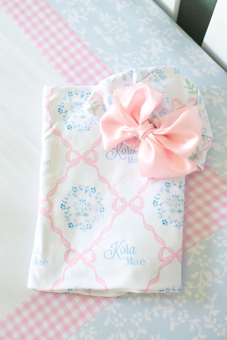 A pink and white baby blanket with a large pink bow and the name "Kora Mae" printed on it, placed on a blue and white checkered bedspread.