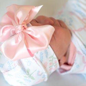 A close-up image of a baby's head wearing a pink bow and a white blanket with a floral pattern.