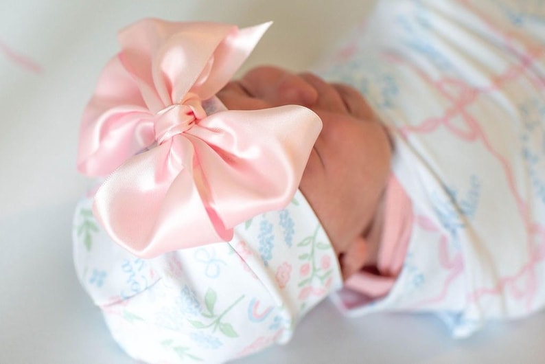 A close-up image of a baby's head wearing a pink bow and a white blanket with a floral pattern.