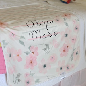 May include: A cream-colored baby blanket with a floral pattern of pink flowers and gray leaves. The name "Aliza Marie" is written in cursive script across the center of the blanket.