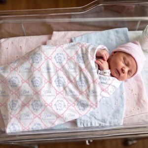 A newborn baby girl is peacefully sleeping in a hospital bed, wrapped in a blanket with the name "Eliza" printed on it.