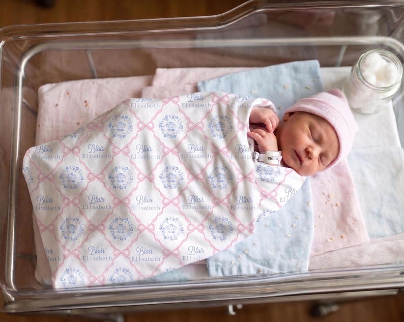 A newborn baby girl is peacefully sleeping in a hospital bed, wrapped in a blanket with the name "Eliza" printed on it.