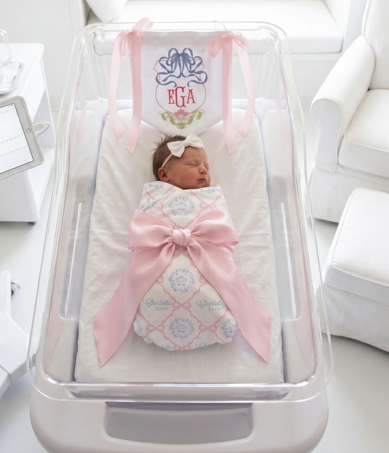A newborn baby girl is peacefully sleeping in a hospital bed, wrapped in a pink blanket and adorned with a pink ribbon.