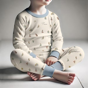 May include: Cream-colored pajama set with a repeating pattern of bunnies and gifts. The long-sleeved top and pants have light blue trim and the name "Finn" printed on them. The child is sitting cross-legged.