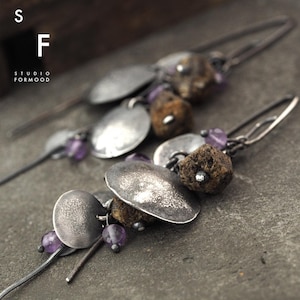 Puede incluir: Un par de pendientes de plata con una estética rústica y orgánica. Cada pendiente presenta un racimo de pequeñas piedras de amatista púrpura, un disco de plata texturizado y una pieza de piedra marrón rugosa. Los pendientes están suspendidos de un fino alambre de plata.