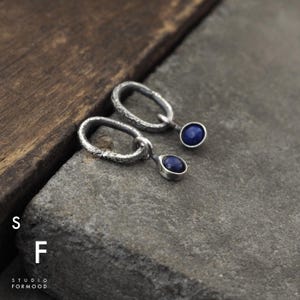 Puede incluir: Un par de pendientes de plata con aros ovalados texturizados y colgantes de piedras preciosas azules.