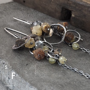 May include: A pair of silver earrings featuring a cluster of amber and clear quartz stones, linked by a silver chain. The earrings are dangling and have a rustic, organic feel.