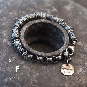 May include: A silver bracelet featuring a chain and a series of small, interconnected rings. The bracelet has a clasp and a small silver charm with the word "formood" on it.