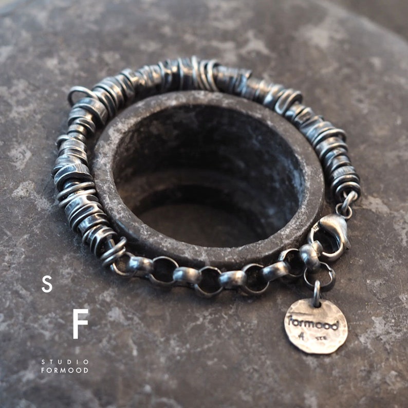 May include: A silver bracelet featuring a chain and a series of small, interconnected rings. The bracelet has a clasp and a small silver charm with the word "formood" on it.