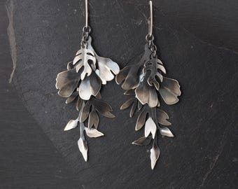 May include: A pair of dangling earrings featuring layered, oxidized silver metal leaves and flower petals. The earrings have a dark, metallic finish with lighter silver highlights on the edges. The text STUDIO FORMOOD is printed at the bottom.