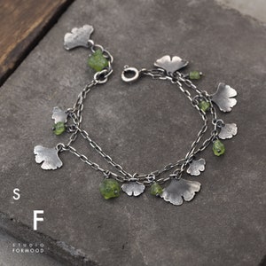 Peut inclure: Un bracelet en argent avec plusieurs breloques en forme de feuilles de ginkgo et des pierres de p&eacute;ridot vertes.