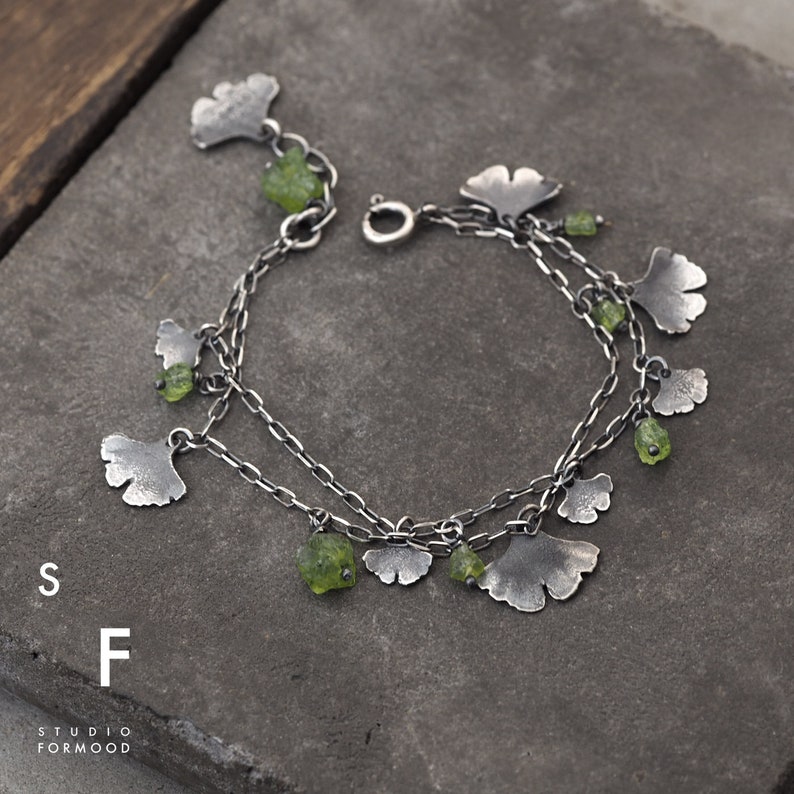 Peut inclure: Un bracelet en argent avec plusieurs breloques en forme de feuilles de ginkgo et des pierres de p&eacute;ridot vertes.