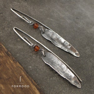Pode incluir: Um par de brincos de prata com formas de lágrima de metal martelado e pequenas contas laranja. Os brincos estão sobre uma superfície cinza texturizada.