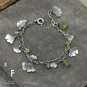 Peut inclure: Un bracelet en argent avec des perles de pierres pr&eacute;cieuses vertes et des breloques en forme de feuilles de ginkgo argent&eacute;es. Le bracelet est dot&eacute; d'un fermoir &agrave; mousqueton.