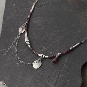 May include: A necklace featuring a strand of small, dark red beads interspersed with silver-toned cylindrical spacers. The piece includes a dangling silver chain with two flat, irregular silver discs and a single faceted dark red gemstone drop.