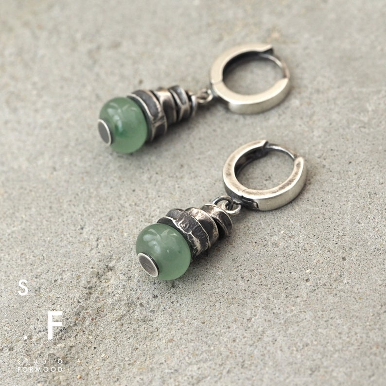 May include: A pair of silver hoop earrings with a green stone pendant. The stone is set in a textured silver setting.