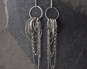 May include: A pair of silver dangle earrings featuring a large circular hoop at the top, from which multiple smaller textured rings and delicate silver chains hang. The earrings are displayed against a dark gray stone surface with the text STUDIO FORMOOD.