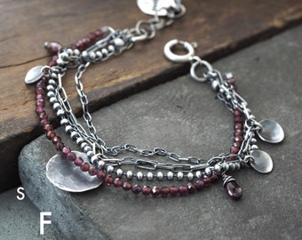 Puede incluir: Un brazalete de múltiples hilos con una cadena de plata, cuentas de plata y cuentas de granate rojo. El brazalete tiene varios abalorios de plata, incluyendo un abalorio redondo con la inscripción "Studio Formwood".