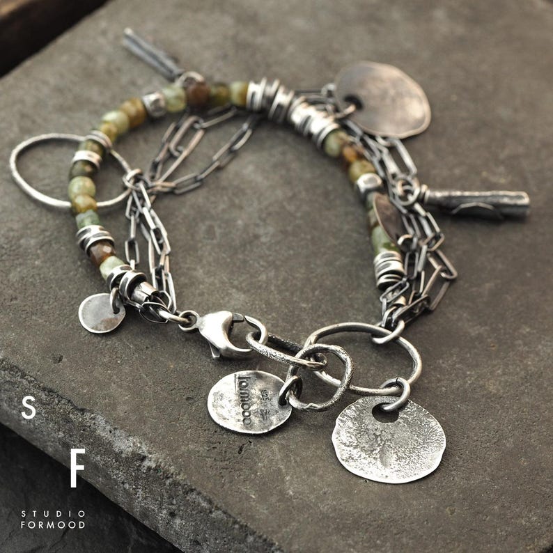 Puede incluir: Una pulsera de plata con m&uacute;ltiples abalorios, incluyendo peque&ntilde;os colgantes redondos y un cierre. La pulsera presenta un hilo de cuentas verdes y marrones y varios eslabones de cadena. El texto "studio formood" es visible.