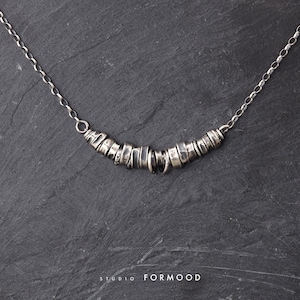 Op de afbeelding: Een zilveren ketting met een fijne schakelketting en een centraal element bestaande uit meerdere kleine, onregelmatige, getextureerde zilveren ringen in een gebogen vorm. De tekst STUDIO FORMOOD staat onderaan afgebeeld.