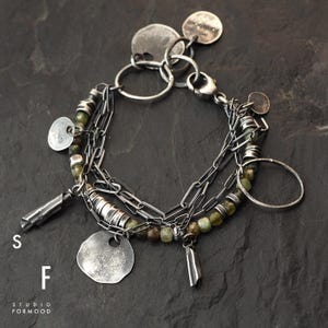Puede incluir: Un brazalete plateado de m&uacute;ltiples hilos con cuentas verdes, colgantes en forma de moneda y elementos cil&iacute;ndricos. El brazalete se muestra sobre una superficie oscura y texturizada. Las letras "S" y "F" y el texto "STUDIO FORMOOD" son visibles.