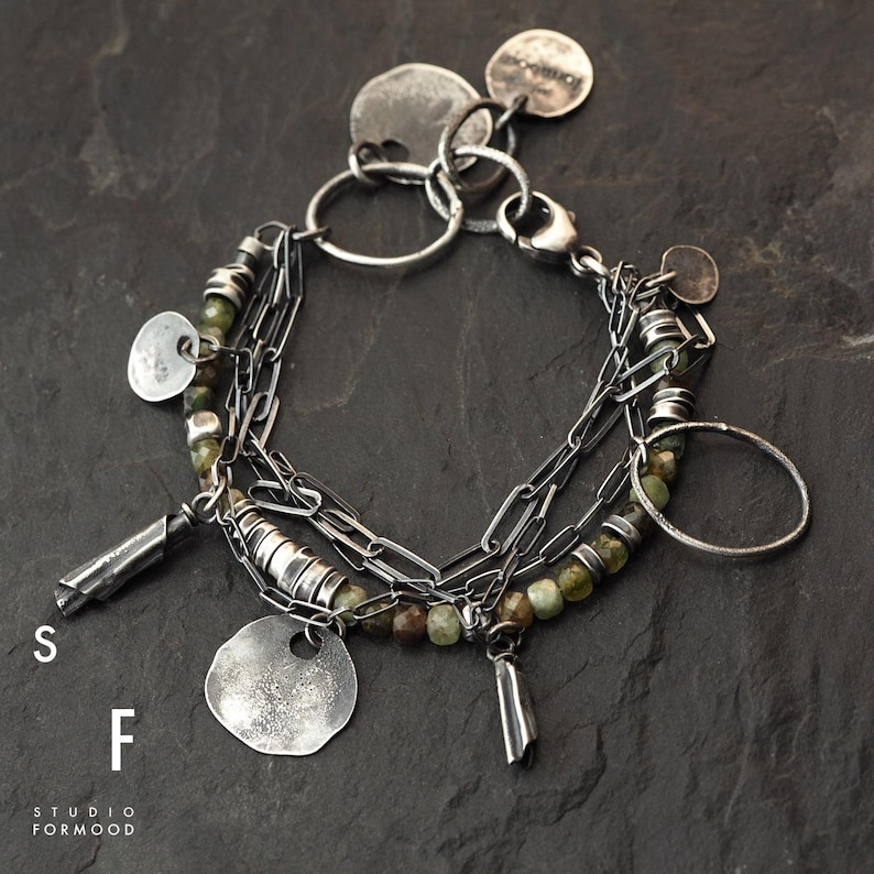 Puede incluir: Un brazalete plateado de m&uacute;ltiples hilos con cuentas verdes, colgantes en forma de moneda y elementos cil&iacute;ndricos. El brazalete se muestra sobre una superficie oscura y texturizada. Las letras "S" y "F" y el texto "STUDIO FORMOOD" son visibles.