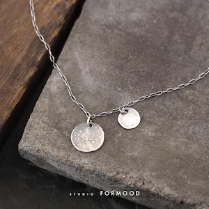 May include: A silver necklace featuring two circular pendants of different sizes. The larger pendant has a textured surface. The necklace is displayed on a gray stone surface. The text "STUDIO FORMOOD" is at the bottom.