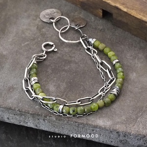 Può includere: Un bracciale a strati con perline di pietre preziose verde oliva e maglie di catena argentate. Il bracciale include una chiusura a moschettone e targhette decorative in metallo. Il testo "STUDIO FORMOOD" è visibile.