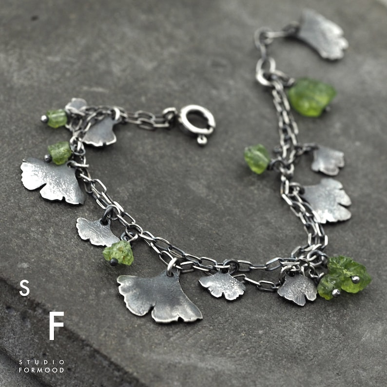 Peut inclure: Un bracelet en argent avec une double rang&eacute;e de maillons. Le bracelet est orn&eacute; de plusieurs breloques en forme de feuilles de ginkgo avec des pierres pr&eacute;cieuses vertes attach&eacute;es. Le bracelet se ferme avec un fermoir &agrave; mousqueton.
