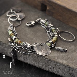 Puede incluir: Una pulsera de plata con m&uacute;ltiples cadenas, con cuentas verde oliva, peque&ntilde;os discos de plata y un colgante martillado texturizado. La pulsera incluye un cierre de mosquet&oacute;n y un dije en forma de llave. El texto "S F STUDIO FORMOOD" es visible.