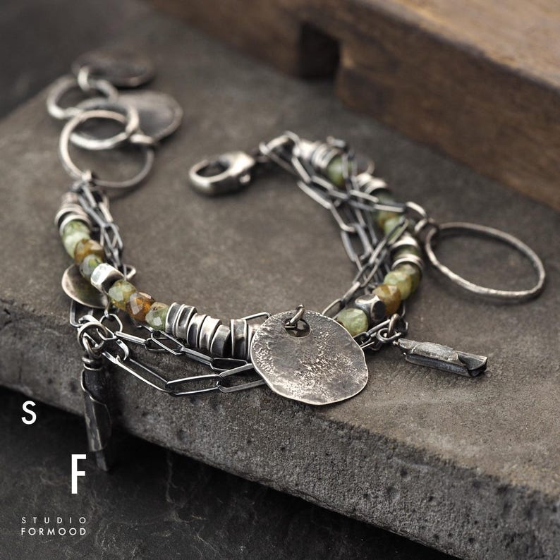 Puede incluir: Una pulsera de plata con m&uacute;ltiples cadenas, con cuentas verde oliva, peque&ntilde;os discos de plata y un colgante martillado texturizado. La pulsera incluye un cierre de mosquet&oacute;n y un dije en forma de llave. El texto "S F STUDIO FORMOOD" es visible.