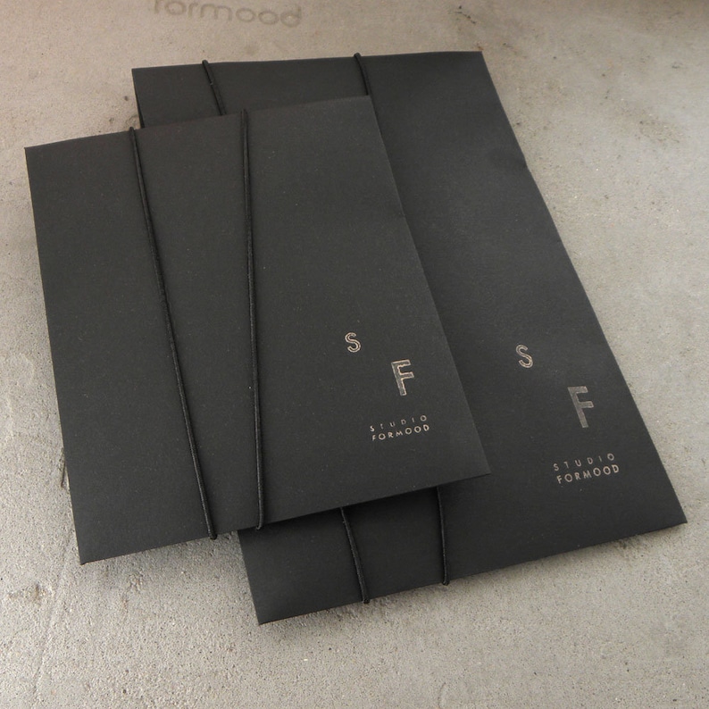 May include: Three black envelopes with black string closures. The envelopes have the text "Studio Formwood" and the letters "SF" printed in silver on the front.