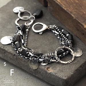 Op de afbeelding: Een meerrijige armband met zwarte kralen, zilveren kettingschakels en zilveren bedels. De armband heeft een sluiting en is gemaakt van metaal.