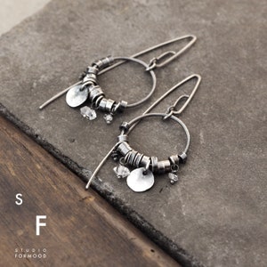 Puede incluir: Un par de pendientes de aro plateados con un diseño único. Cada pendiente presenta un aro circular adornado con pequeñas cuentas plateadas, un disco plateado plano y cristales transparentes colgantes. Los pendientes se exhiben sobre una superficie gris.