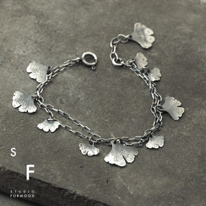 May include: A silver bracelet with a delicate chain and multiple ginkgo leaf charms. The leaves have a textured surface and are connected to the chain with small loops.