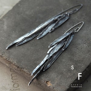 Puede incluir: Un par de pendientes largos de plata con un diseño texturizado y orgánico. Los pendientes están hechos de múltiples piezas delgadas de metal superpuestas, ligeramente curvadas y con un acabado rugoso y martillado. Los pendientes están suspendidos de un simple gancho.