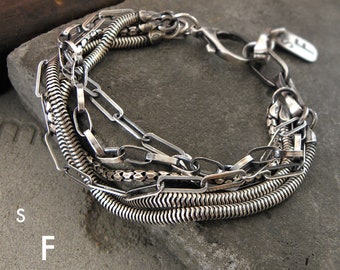 Puede incluir: Un brazalete de plata con múltiples eslabones de cadena, incluyendo una cadena de serpiente, una cadena de caja y una cadena de bordillo. El brazalete tiene un cierre de broche y una pequeña etiqueta con la letra "F" en ella.