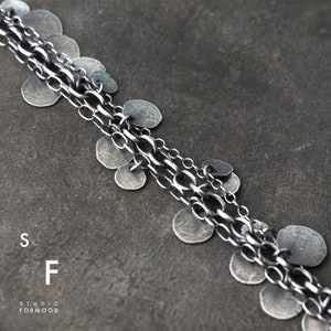 May include: A silver chain necklace with multiple strands and small, round silver charms. The necklace is on a gray background.