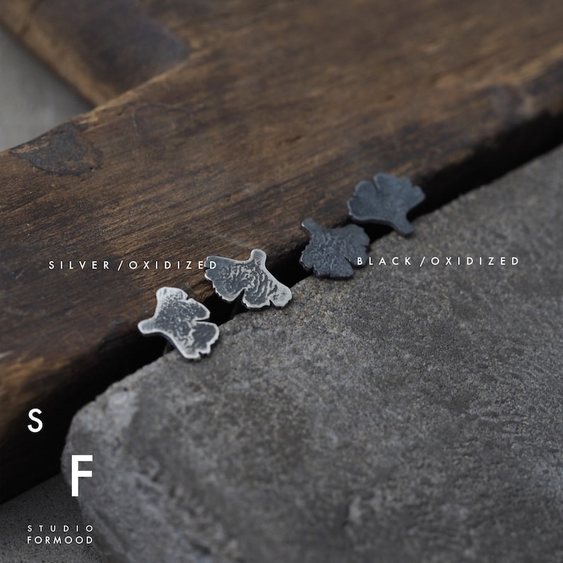 May include: Two pairs of oxidized silver earrings in the shape of a ginkgo leaf. The earrings are displayed on a wooden surface. The text "SILVER/OXIDIZED" and "BLACK/OXIDIZED" is visible below the earrings.