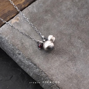 Op de afbeelding: Een zilverkleurige ketting met twee bolvormige bedels met een matte afwerking en een kleine, donkerrode kraal. De ketting wordt getoond met de tekst STUDIO FORMOOD in het wit onderaan de afbeelding.