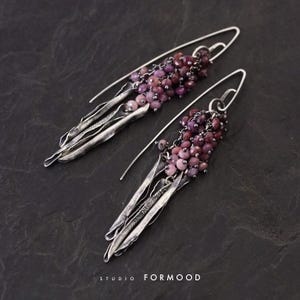 Può includere: Orecchini in argento con un gruppo di piccole perline rotonde viola e lunghi pendenti argentati a forma di foglia strutturata. Gli orecchini hanno un design a gancio. Le parole "studio FORMOOD" sono in basso.