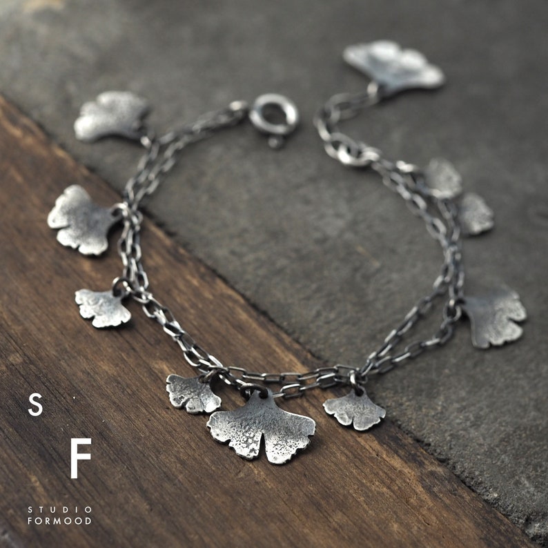 May include: A silver bracelet with a delicate chain and multiple ginkgo leaf charms. The bracelet is on a wooden surface.