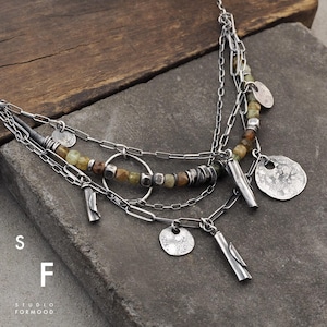 May include: A silver necklace with multiple strands, featuring small green and brown beads, a circle pendant, and several silver charms. The necklace is displayed on a rustic wooden surface.