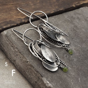 May include: Silver earrings with multiple layered circles and oval shapes, accented with small green gemstones. The earrings are hanging from a silver hook.