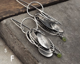 May include: Silver earrings with multiple layered circles and oval shapes, accented with small green gemstones. The earrings are hanging from a silver hook.