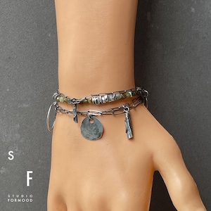 Puede incluir: Una pulsera de plata en capas con m&uacute;ltiples dijes, incluyendo un c&iacute;rculo, una cruz y un disco. La pulsera tambi&eacute;n presenta un dise&ntilde;o de eslabones de cadena y peque&ntilde;as cuentas verdes. Las letras "S" y "F" son visibles, junto con "STUDIO FORMOOD".