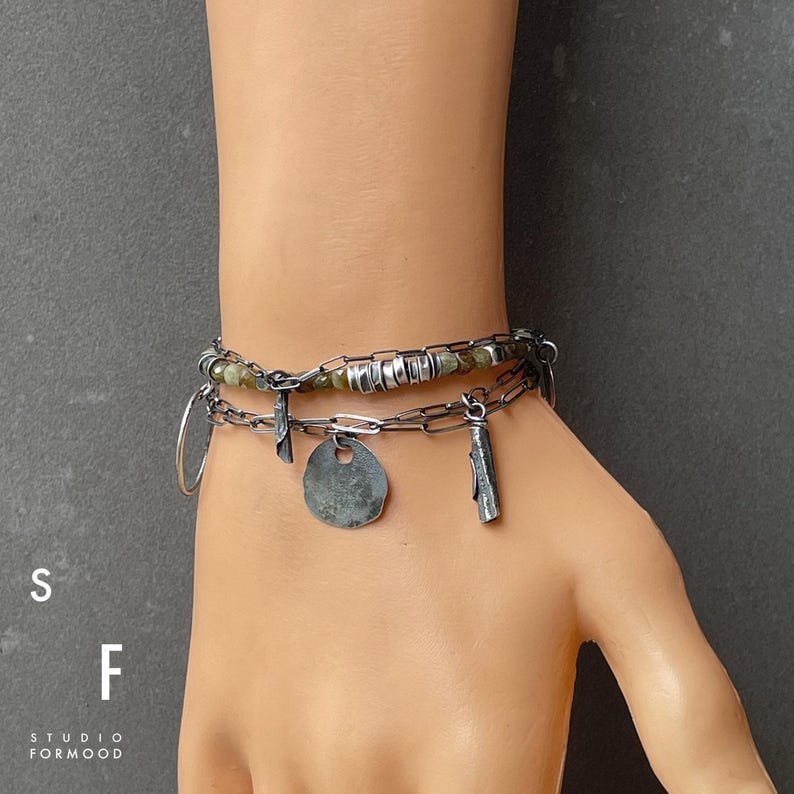 Puede incluir: Una pulsera de plata en capas con m&uacute;ltiples dijes, incluyendo un c&iacute;rculo, una cruz y un disco. La pulsera tambi&eacute;n presenta un dise&ntilde;o de eslabones de cadena y peque&ntilde;as cuentas verdes. Las letras "S" y "F" son visibles, junto con "STUDIO FORMOOD".
