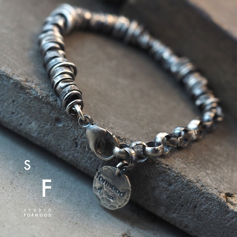 May include: Silver bracelet featuring a chain and a series of stacked rings. The bracelet has a clasp and a small silver tag with the word "formmood" on it.