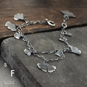 May include: A silver bracelet with a delicate chain and multiple ginkgo leaf charms. The bracelet is on a wooden surface.