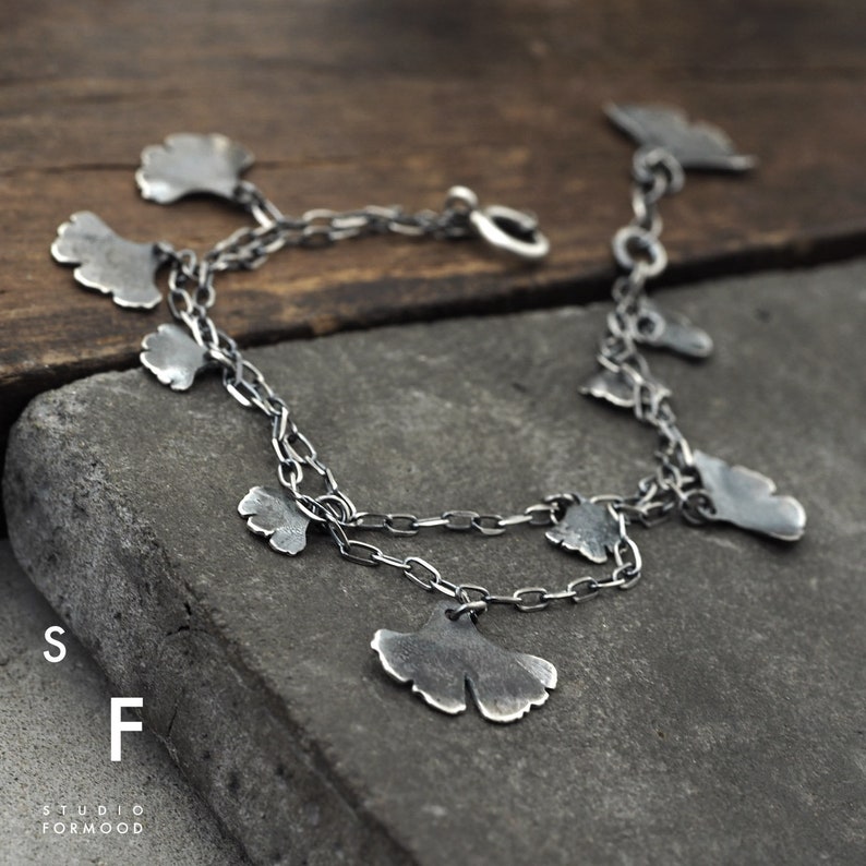 May include: A silver bracelet with a delicate chain and multiple ginkgo leaf charms. The bracelet is on a wooden surface.